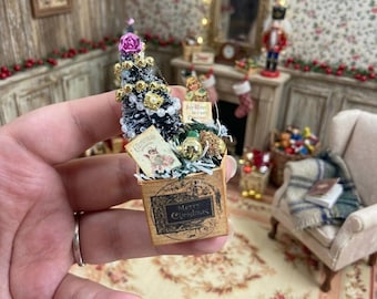 May include: A miniature Christmas tree in a square cardboard box with the text Merry Christmas. The tree is decorated with gold beads, white pearls, and a pink flower. A small rectangular card with a vintage illustration is tucked into the box.