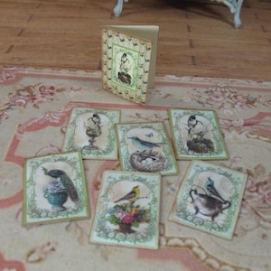 Può includere: Una collezione di biglietti di auguri in miniatura a tema uccelli e un biglietto pieghevole abbinato. I biglietti presentano illustrazioni in stile vintage di vari uccelli, fiori ed elementi decorativi all'interno di un bordo verde. I biglietti sono disposti su una superficie a motivi.