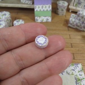 May include: A small, round, white tin with a lavender design and the text "Lavender" on it. The tin is being held in a hand.