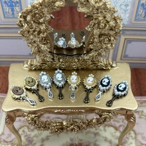 Può includere: Una collezione di specchietti da mano in miniatura con disegni a cammeo, disposti su un tavolo da toeletta dorato. Gli specchietti presentano cornici ornate, dettagli di perle e vari colori cammeo. Un grande specchio decorativo è sullo sfondo.
