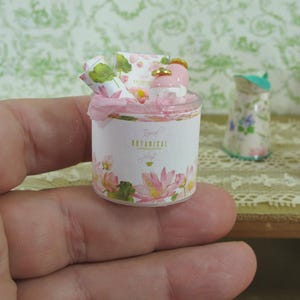 Puede incluir: Miniatura de un juego de regalo floral. El juego incluye un pequeño recipiente cilíndrico con el texto "Tropical Botanical" e ilustraciones de flores de loto rosas. El recipiente contiene una botella pequeña, papel enrollado y papel de seda rosa. Otros artículos florales en miniatura están en el fondo.