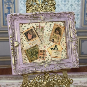 May include: A square lavender picture frame with gold-painted ornate edges, featuring a collage of vintage-style paper scraps, a lace strip, and small illustrations of a person, a butterfly, and text including How to Make Dresses.