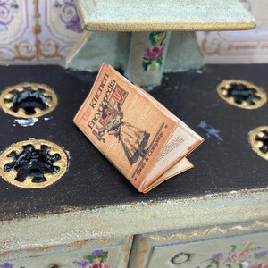 May include: A miniature book titled "The Kitchen Encyclopedia" with a vintage illustration on the cover. The book is open, revealing its pages. It rests on a black surface with gold accents, suggesting a kitchen or dollhouse setting.