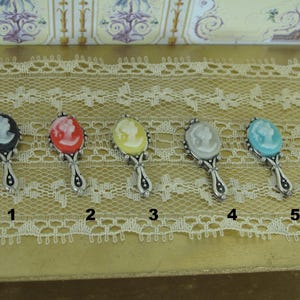 May include: Five miniature hand mirrors with different colored cameo portraits. The mirrors are arranged in a row on a white lace surface. The mirrors are numbered 1 through 5.