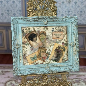 May include: A light blue ornate frame with gold highlights containing a collage of vintage-style paper ephemera, including a portrait of a woman, a butterfly, an Eiffel Tower illustration, and a label with the text How to Make Dresses.