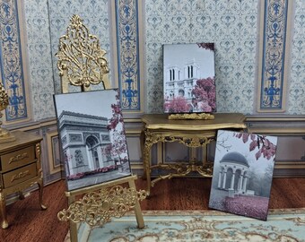 May include: Miniature gold easel holding a black and white print of the Arc de Triomphe in Paris, France. Two other prints depict the Notre Dame Cathedral and a gazebo. The prints have pink accents and are displayed on a gold table.