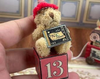 May include: A miniature teddy bear wearing a red party hat and holding a small sign that reads "ABC Primer 123." The bear is sitting on a red block with the number 13. The bear is light brown with a dark brown nose.