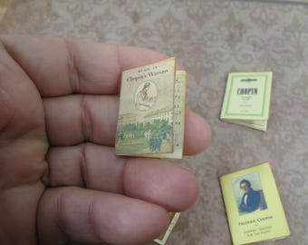 May include: A miniature book titled "Music in Chopin's Warsaw" with a picture of a person on the cover. The book is held in a hand. There are two other miniature books in the background, one titled "Chopin" and the other titled "Frederic Chopin - Chopin's Waltzes for the Piano".
