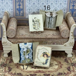 Può includere: Miniature per casa delle bambole, tra cui un vestito grigio con finiture in pizzo bianco, un vestito blu con pizzo bianco e due cartoline in stile vintage. Gli articoli sono esposti su una panca decorativa e un tappeto floreale.