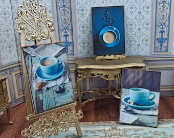May include: Miniature art prints of coffee cups on easels and a table. The prints feature blue coffee cups on saucers, with a spoon and a cookie. The background includes a blue and white patterned wall and a gold-colored table and easel.
