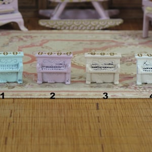 Könnte beinhalten: Vier Miniaturklaviere in verschiedenen Farben: hellblau, Lavendel, weiß und hellblau mit goldenen Akzenten. Die Klaviere sind in einer Reihe auf einem Teppich angeordnet, wobei sich unter jedem Klavier die Zahlen 1 bis 4 befinden.