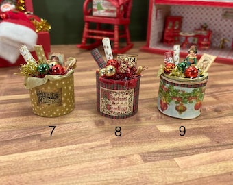 May include: Three miniature Christmas gift boxes. The boxes are cylindrical and decorated with festive patterns and embellishments. Each box contains small ornaments, decorative paper, and other holiday-themed items. The boxes are numbered 7, 8, and 9.
