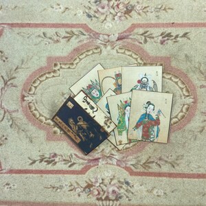 May include: A set of six vintage Chinese opera playing cards featuring colorful illustrations of characters in traditional costumes. The cards are arranged on a beige rug with a floral pattern.