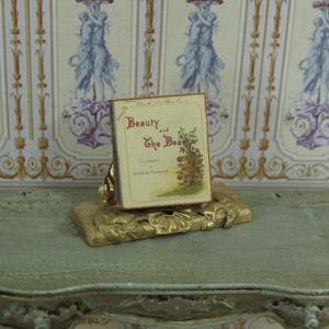 Peut inclure: Un support décoratif doré avec un livre intitulé "La Belle et la Bête" dessus. Le livre a un motif floral avec des roses rouges sur fond blanc.