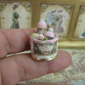 Peut inclure: Miniature ensemble de cosmétiques roses et blancs dans un récipient rond avec un motif floral. L'ensemble comprend deux flacons et une petite carte, avec un bord doré. Le récipient est rempli de petites fleurs.