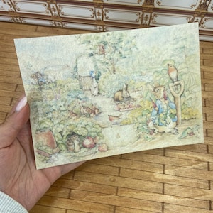 May include: A vintage-style postcard featuring a watercolor illustration of Peter Rabbit and other characters in a garden setting. The artwork depicts rabbits, birds, and a gardener amidst lush greenery, with soft pastel colors.