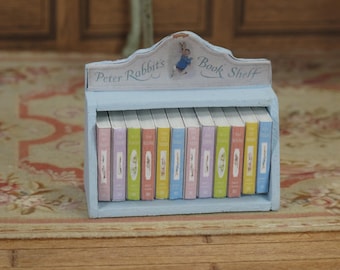 Könnte beinhalten: Ein hellblaues Miniatur-Bücherregal mit der Aufschrift "Peter Rabbit's Book Shelf" auf der Vorderseite. Das Regal ist mit Miniaturbüchern mit bunten Umschlägen gefüllt.