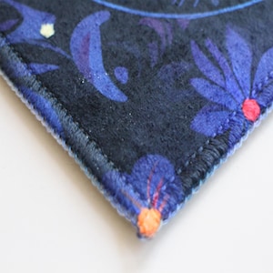 Pu&ograve; includere: Primo piano del bordo di un tessuto blu scuro con un motivo floreale. Il tessuto presenta un motivo floreale blu, viola e arancione. Il bordo del tessuto &egrave; cucito con un filo blu chiaro.