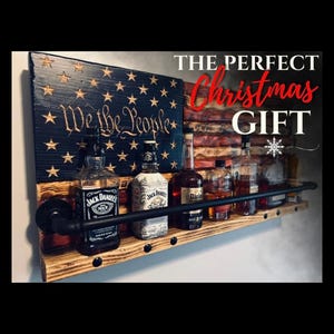 May include: A wall-mounted wooden liquor shelf featuring a charred American flag design with the text We the People. The shelf includes a black metal pipe railing and holds five glass bottles of various spirits. Text reads THE PERFECT Christmas GIFT.