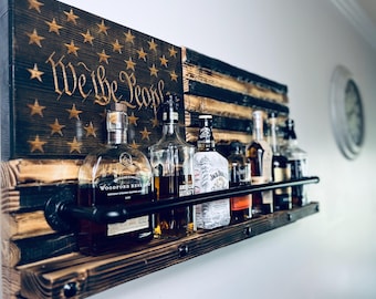 Könnte beinhalten: Ein rustikales Barregal aus Holz mit einem verwitterten Design der amerikanischen Flagge. Das Regal zeigt verschiedene Spirituosenflaschen und trägt den Text "We the People" in goldfarbenen Buchstaben. Eine schwarze Metallstange ist installiert.