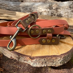 Puede incluir: Una correa para perros enrollada, de color óxido, con detalles de cuero marrón a juego y herrajes de color latón. La correa está enrollada sobre una superficie de madera, mostrando su diseño. Incluye un mosquetón y un anillo.