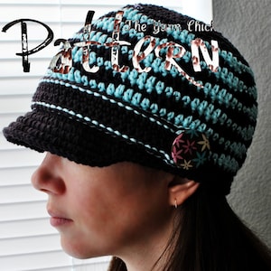 May include: A brown and blue crocheted hat with a brim. The hat has a button with a floral design on the side. The text "The Yarn Chick" is visible on the hat.