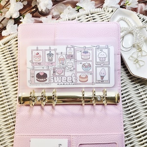 Op de afbeelding: Een lichtroze binder met gouden ringen, open om een ​​transparante zak te onthullen met een kaart met illustraties van desserts en de woorden "Sweet moments". De kaart bevat tekeningen van cupcakes en cakes. De binder ligt op een geweven oppervlak.