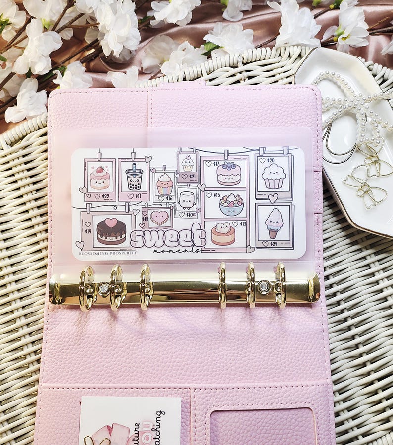Op de afbeelding: Een lichtroze binder met gouden ringen, open om een ​​transparante zak te onthullen met een kaart met illustraties van desserts en de woorden "Sweet moments". De kaart bevat tekeningen van cupcakes en cakes. De binder ligt op een geweven oppervlak.