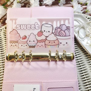 Op de afbeelding: Een lichtroze binder met goudkleurige ringen, open om een ​​transparante zak met het woord "sweet" en illustraties van cupcakes en desserts te onthullen. De binder staat op een wit geweven oppervlak, met een decoratief dienblad en sieraden op de achtergrond.