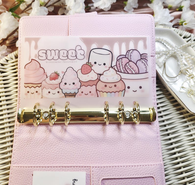 Op de afbeelding: Een lichtroze binder met goudkleurige ringen, open om een ​​transparante zak met het woord "sweet" en illustraties van cupcakes en desserts te onthullen. De binder staat op een wit geweven oppervlak, met een decoratief dienblad en sieraden op de achtergrond.