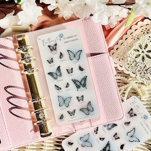 Puede incluir: Una agenda rosa con detalles dorados contiene una lámina transparente de pegatinas de mariposas. Las pegatinas presentan varios diseños de mariposas con etiquetas de precios. El texto de la lámina dice "The Butterfly Life is a reminder that chance is beautiful."