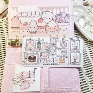 Op de afbeelding: Een roze planner met decoratieve inzetstukken met cartoon desserts zoals cupcakes en macarons. De inzetstukken hebben de woorden "sweet" en "sweet moments" erop gedrukt. Een kleine kaart met een bloem en de tekst "future is watching" is ook zichtbaar.