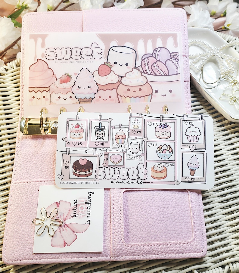 Op de afbeelding: Een roze planner met decoratieve inzetstukken met cartoon desserts zoals cupcakes en macarons. De inzetstukken hebben de woorden "sweet" en "sweet moments" erop gedrukt. Een kleine kaart met een bloem en de tekst "future is watching" is ook zichtbaar.