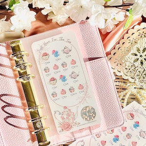 May include: A pink planner with gold accents, open to reveal decorative stickers. The stickers feature teacups, cupcakes, and floral designs with the text "Just Here For The Tea" and "Blossoming Prosperity".