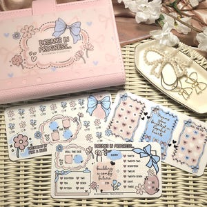 May include: A pink planner and decorative stickers with floral and bow designs. The planner has the text "DREAMS IN PROGRESS..." and the stickers have phrases like "ROLL THE DICE" and "building a comfy future". A white tray with bow-shaped jewelry is also present.