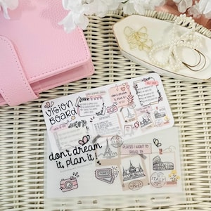 Puede incluir: Una agenda rosa con una inserción de tablero de visión con ilustraciones de temática de viajes y el texto "don't dream it, plan it!". Un plato pequeño contiene clips de papel dorados en forma de lazo y un collar de perlas. El fondo incluye flores blancas.
