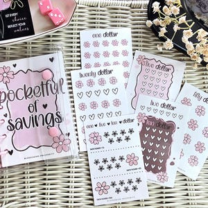 May include: A clear plastic binder with pink rings and a cover labeled pocketful of savings, accompanied by several white paper inserts featuring pink flower and heart icons labeled with various dollar amounts for financial tracking.