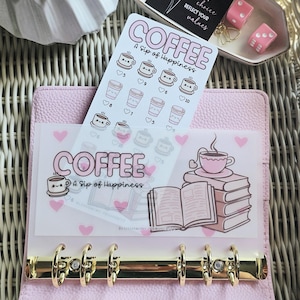 May include: A light pink six-ring binder containing a translucent dashboard with a coffee and book illustration and the text "COFFEE A Sip of Happiness," alongside a sticker sheet featuring coffee cup icons and heart-shaped tracking numbers.