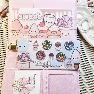 Op de afbeelding: Roze planner met stickers met cupcakes, macarons en het woord "sweet". De stickers hebben holografische accenten en de zin "life is sweet, enjoy the treat". Een kaart met een bloem en de tekst "future is watching" is ook zichtbaar.