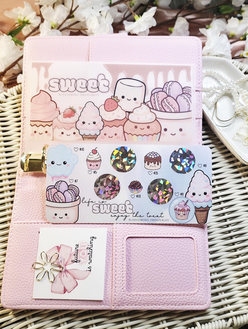 Op de afbeelding: Roze planner met stickers met cupcakes, macarons en het woord "sweet". De stickers hebben holografische accenten en de zin "life is sweet, enjoy the treat". Een kaart met een bloem en de tekst "future is watching" is ook zichtbaar.