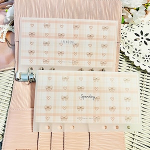 May include: Two clear plastic budget envelopes with a pink plaid pattern, bows, and hearts. The word "Spending" is written on each envelope. The envelopes are inside a pink binder.