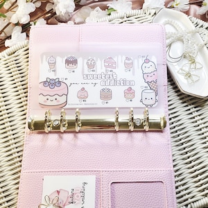 Op de afbeelding: Een roze planner in binderstijl van imitatieleer met goudkleurige ringen. Een doorzichtige plastic hoes aan de binnenkant toont ge&iuml;llustreerde desserts en de tekst "Sweetest Addiction". Een kleine kaart met tekst is zichtbaar in een vak.