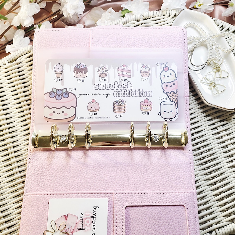 Op de afbeelding: Een roze planner in binderstijl van imitatieleer met goudkleurige ringen. Een doorzichtige plastic hoes aan de binnenkant toont ge&iuml;llustreerde desserts en de tekst "Sweetest Addiction". Een kleine kaart met tekst is zichtbaar in een vak.