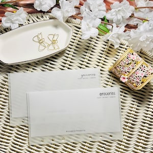 May include: Two gold bow-shaped paper clips rest on a white rectangular dish with a black trim. Two white envelopes with the word "groceries" are on a woven surface. A decorative box and white flowers are also present.