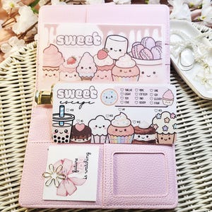 Op de afbeelding: Een roze planner met een gouden sluiting, opengeklapt om decoratieve kaarten met cupcake-illustraties en de woorden "Sweet" en "Sweet Escape" te onthullen. Een kleine kaart met een bloemenontwerp en de tekst "future is watching" is ook zichtbaar.