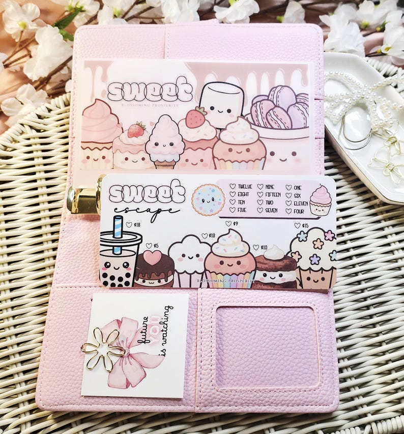 Op de afbeelding: Een roze planner met een gouden sluiting, opengeklapt om decoratieve kaarten met cupcake-illustraties en de woorden "Sweet" en "Sweet Escape" te onthullen. Een kleine kaart met een bloemenontwerp en de tekst "future is watching" is ook zichtbaar.