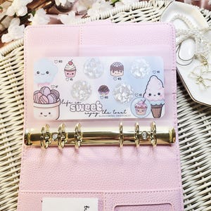 Op de afbeelding: Een roze planner met gouden ringen, open om een ​​transparante zak met stickers met dessertthema's te laten zien. De stickers bevatten cupcakes, ijsjes en macarons. De tekst "life is sweet, enjoy the treat" is ook zichtbaar.