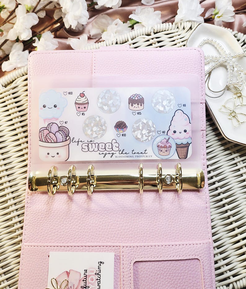 Op de afbeelding: Een roze planner met gouden ringen, open om een ​​transparante zak met stickers met dessertthema's te laten zien. De stickers bevatten cupcakes, ijsjes en macarons. De tekst "life is sweet, enjoy the treat" is ook zichtbaar.