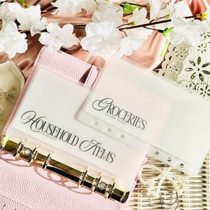 Könnte beinhalten: Ein rosa Ordner mit goldenen Akzenten enthält transparente Kunststoffeinsätze mit der Aufschrift "Household Items" und "Groceries". Weiße Blumen und goldene Schleifenornamente im Hintergrund deuten auf ein Organisationsthema hin.