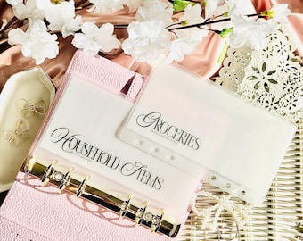 May include: A pink binder with gold detailing contains clear plastic inserts labelled "Household Items" and "Groceries." White flowers and gold bow ornaments are in the background, suggesting an organisational theme.